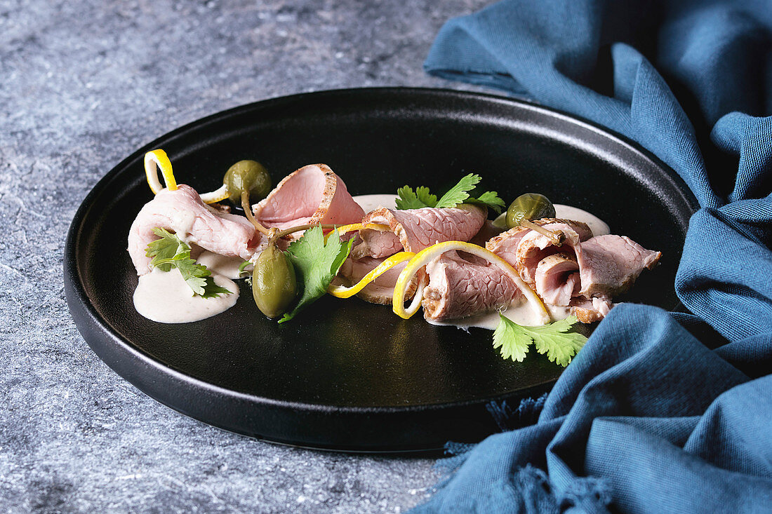 Vitello tonnato italian dish. Thin sliced veal with tuna sauce, capers and coriander served on black plate with blue textile