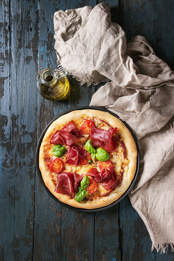 Traditional pizza with bresaola, cheese, tomatoes and basil served on black plate with textile and olive oil over dark blue wooden background