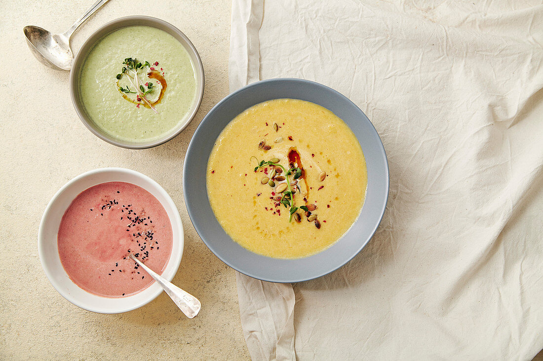 Three bowls with colorful vegetarian creamy soups with broccoli, pumpkin and beetroot garnished with pumpkin seeds and olive oil