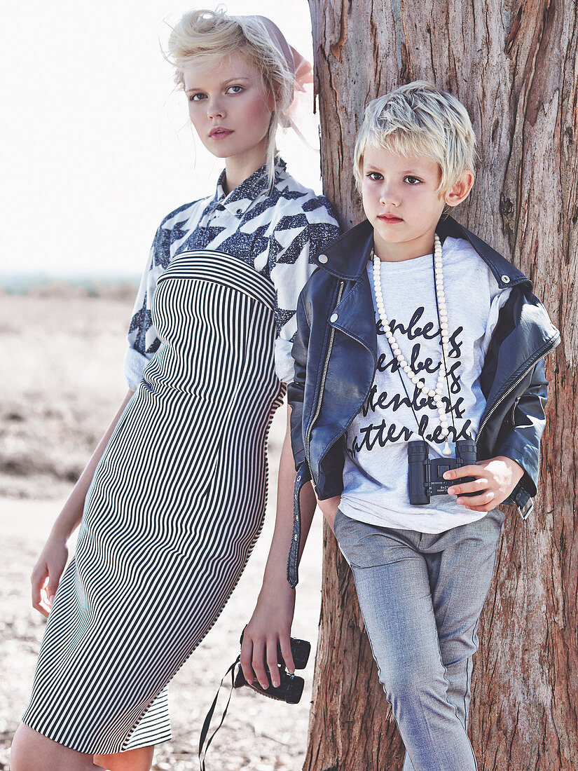 A blonde woman wearing a striped dress leaning against a tree with a blond boy