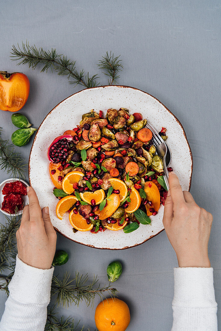A woman serving a bowl of roasted brussels sprouts salad with carrot, orange, persimmon and pomegranate