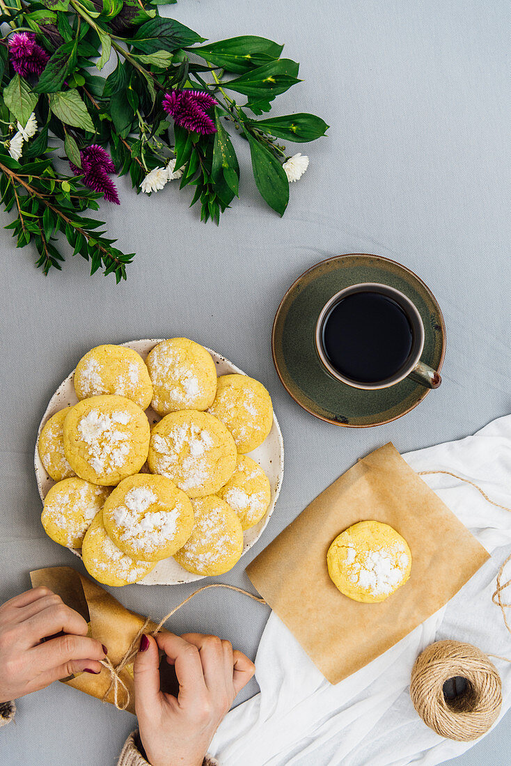 A woman wrapping lemon cookies and more lemon cookies stacked on a plate, a cup of coffee and flowers