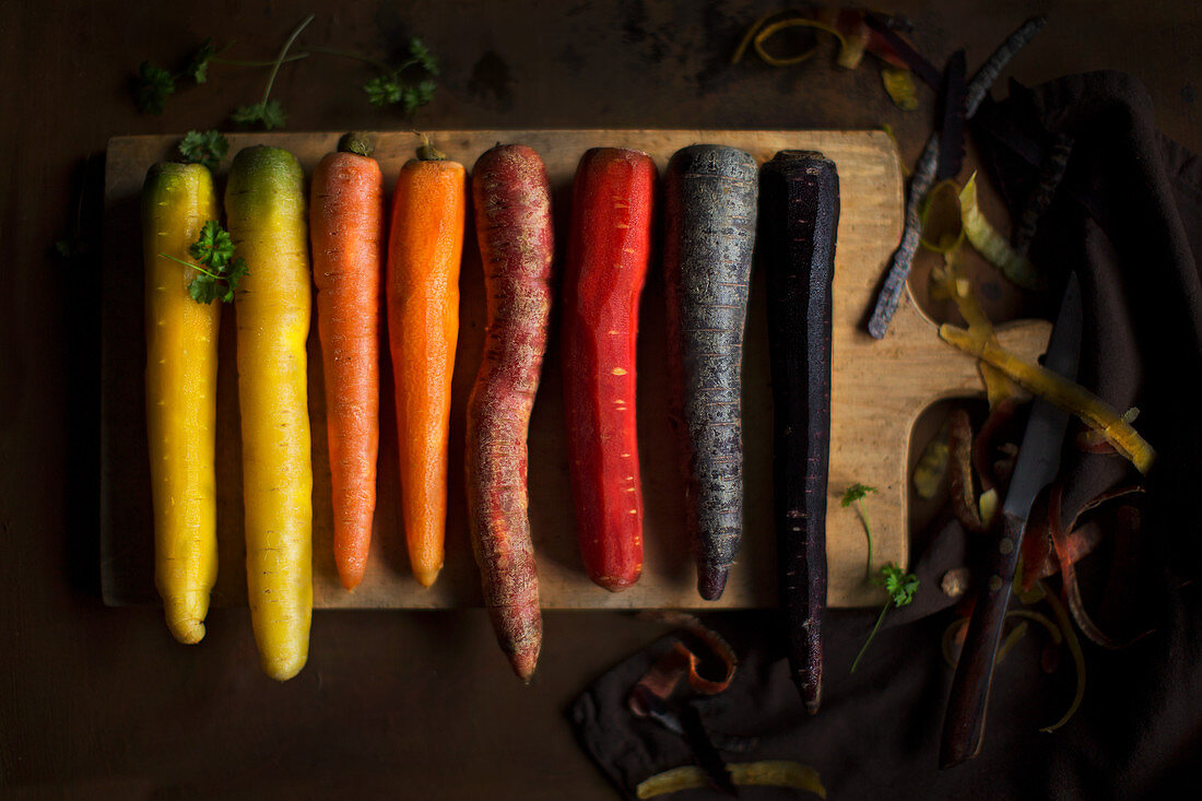 Four different carrots (yellow, orange, red and purple) partially peeled on a wooden board with a peeler and a napkin