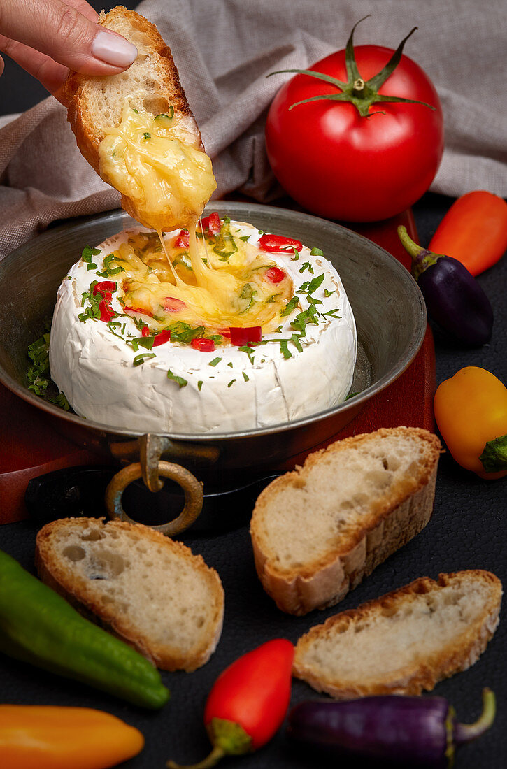 Woman hand dipping slice of bread inside melted Camembert cheese, seasoned with herbs and peppers