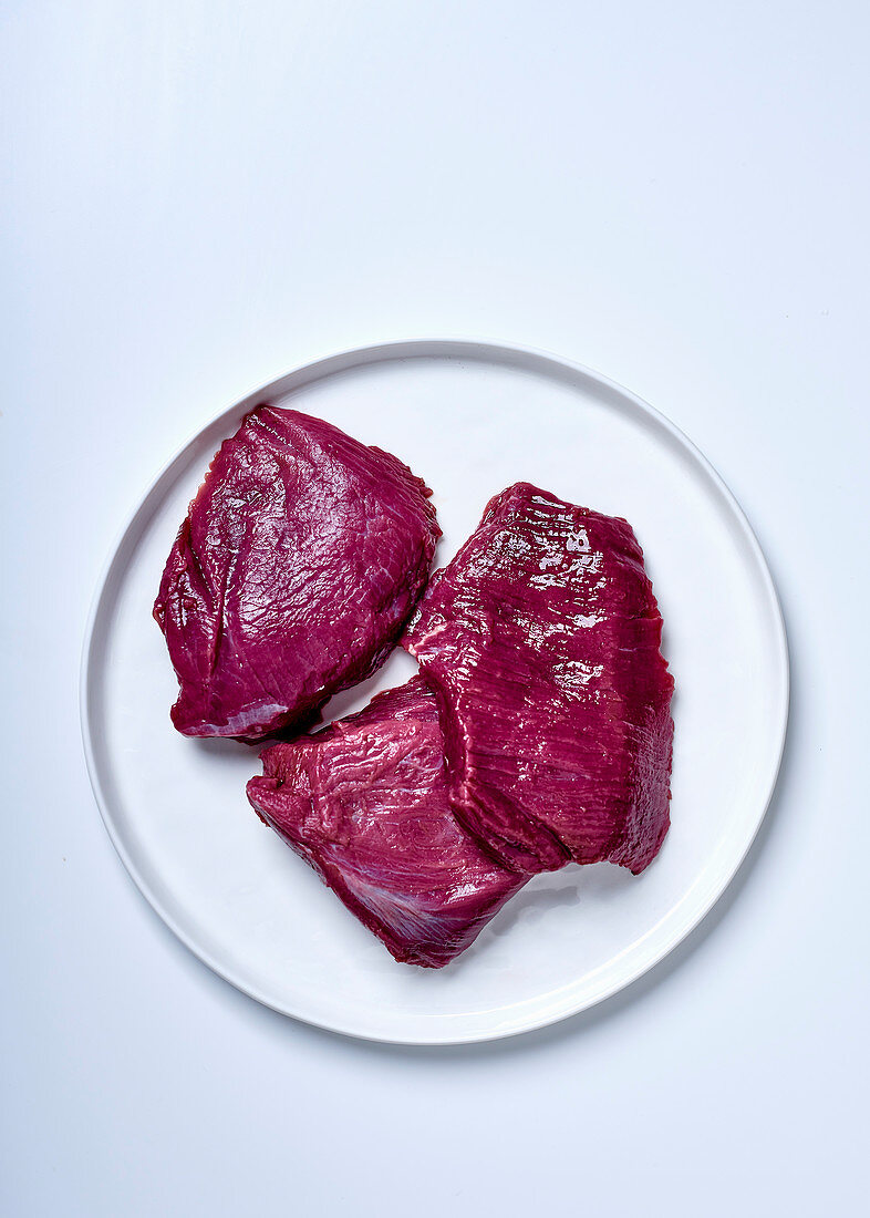 Three raw ostrich steaks on a plate against a white background (top view)