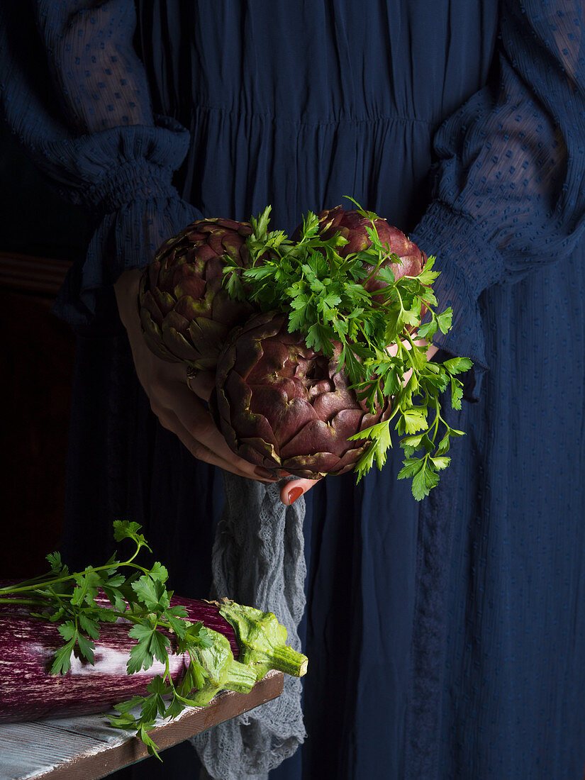 Woman in blue dress holding a bunch of artichokes with parsley and aubergines on the table