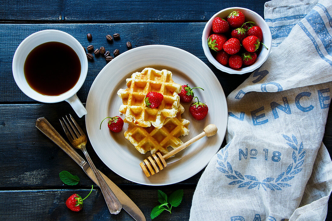 Breakfast belgian waffles stacked on a plate, served with coffee, fresh strawberries and mint at textile napkin over old wooden table