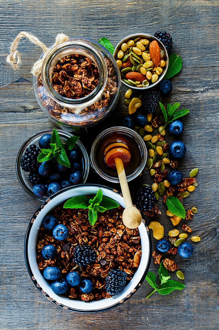Top view of chocolate granola in open jar and honey served with berries, nuts and fresh mint for tasty breakfast on rustin wooden table