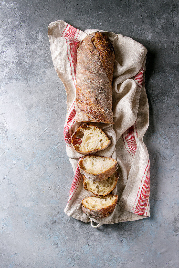 Sliced fresh baked artisan whole grain ciabatta bread on kitchen towel over grey texture background