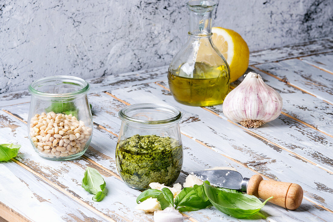 Basil pesto sauce in glass jar with fresh basil, olive oil, parmesan cheese, garlic, pine nuts, lemon on wooden kitchen table