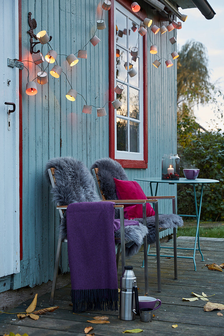 DIY-Lichterkette aus Papierbackförmchen, Plaid und Schaffelle auf Gartenstühlen, Frau im Hintergrund