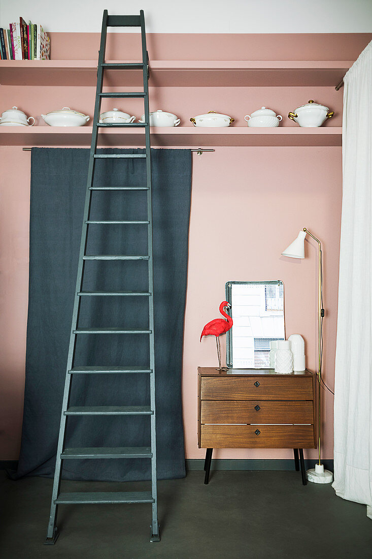 Standard lamp, chest of drawers and length of green fabric below china on shelf and ladder against pink wall