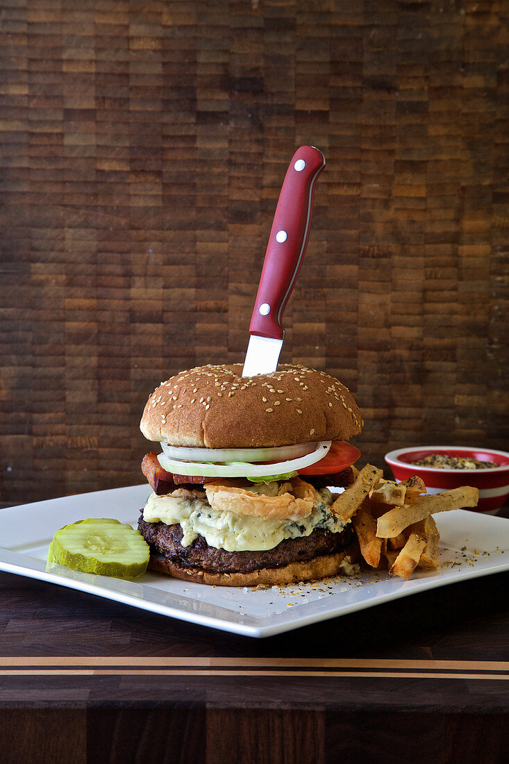 Burger with blue cheese, lettuce, deep fried onions, fries, pickle and tomato on a dark rustic background with a knife