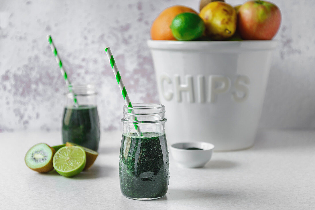 Spirulina and green fruit smoothie in glass jars, halved kiwi and limes, a big white bowl of assorted fruit on a light colored tabletop