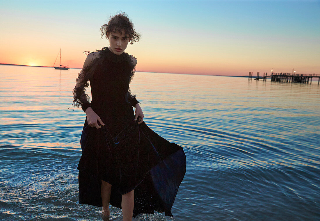 A dark-haired woman wearing a long evening dress by the sea at sunset