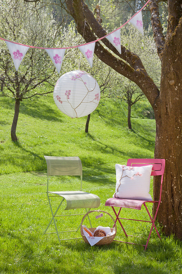 Romantic seat below lantern and bunting on flowering cherry tree:handmade cushion on chair next to branch wound with white yarn and decorated with crocheted flowers