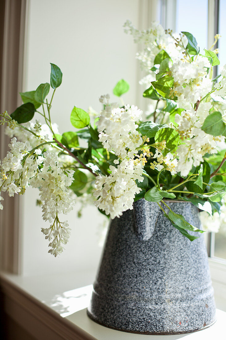 Arrangement of white lilac in old enamel jug on windowsill