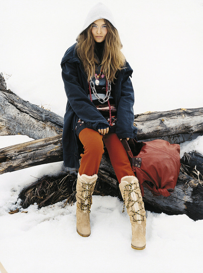 A young woman in the snow wearing a dark coat, rust-brown trousers and boots
