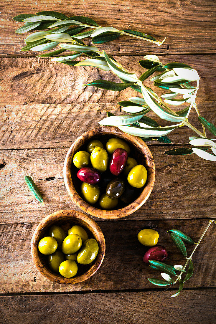 Various olives in a wooden bowl next to olive sprigs (seen from above)