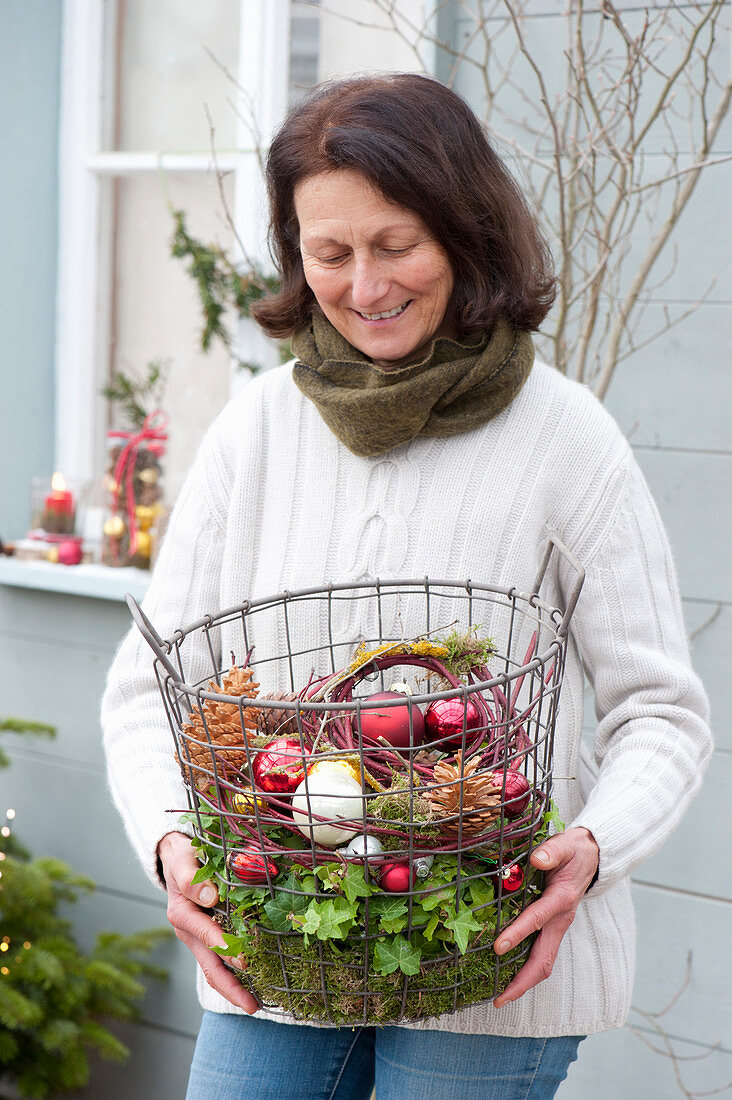 Woman bringing wire basket with Christmas baubles, cones and moss