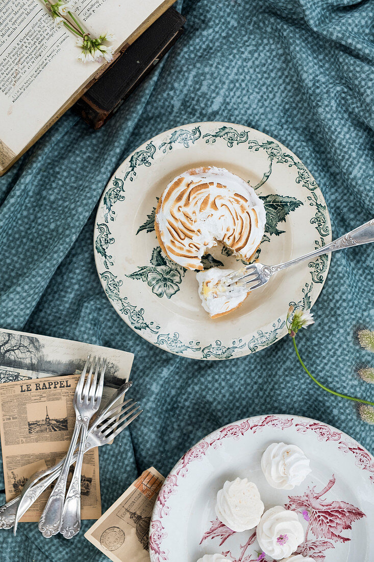 A brocante plate with a citron meringue and little meringues on a plate with old postcards on a cloth