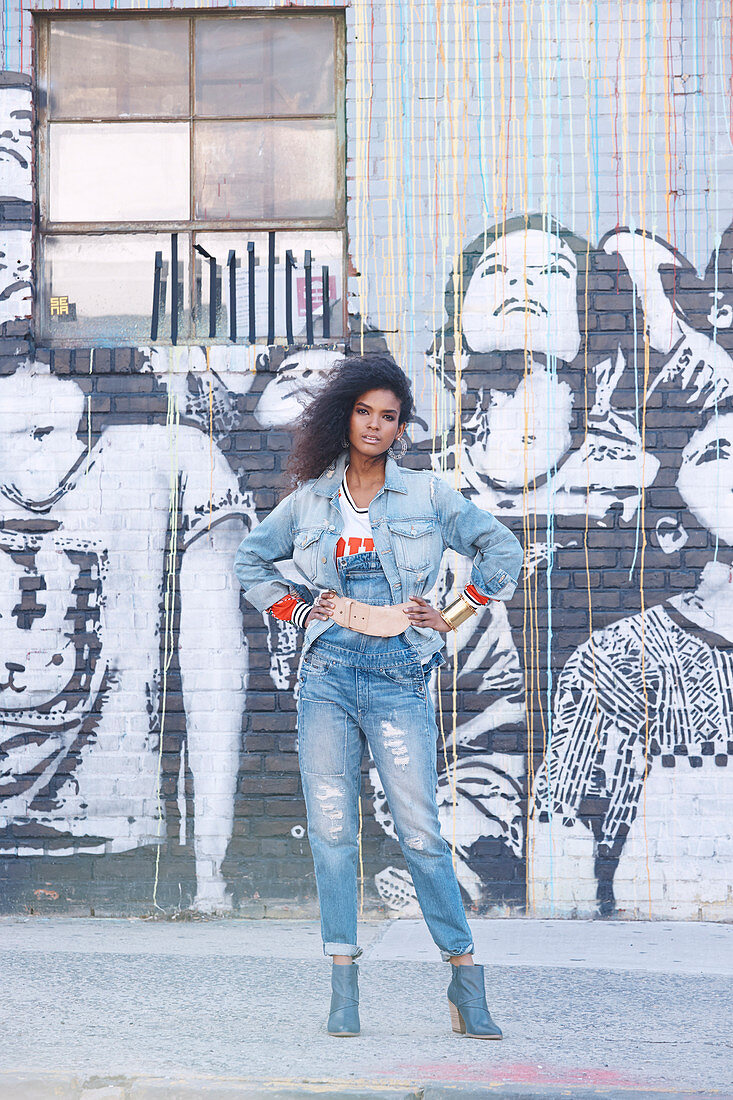 A dark-haired woman wearing denim dungarees and a denim jacket standing against a graffitied wall
