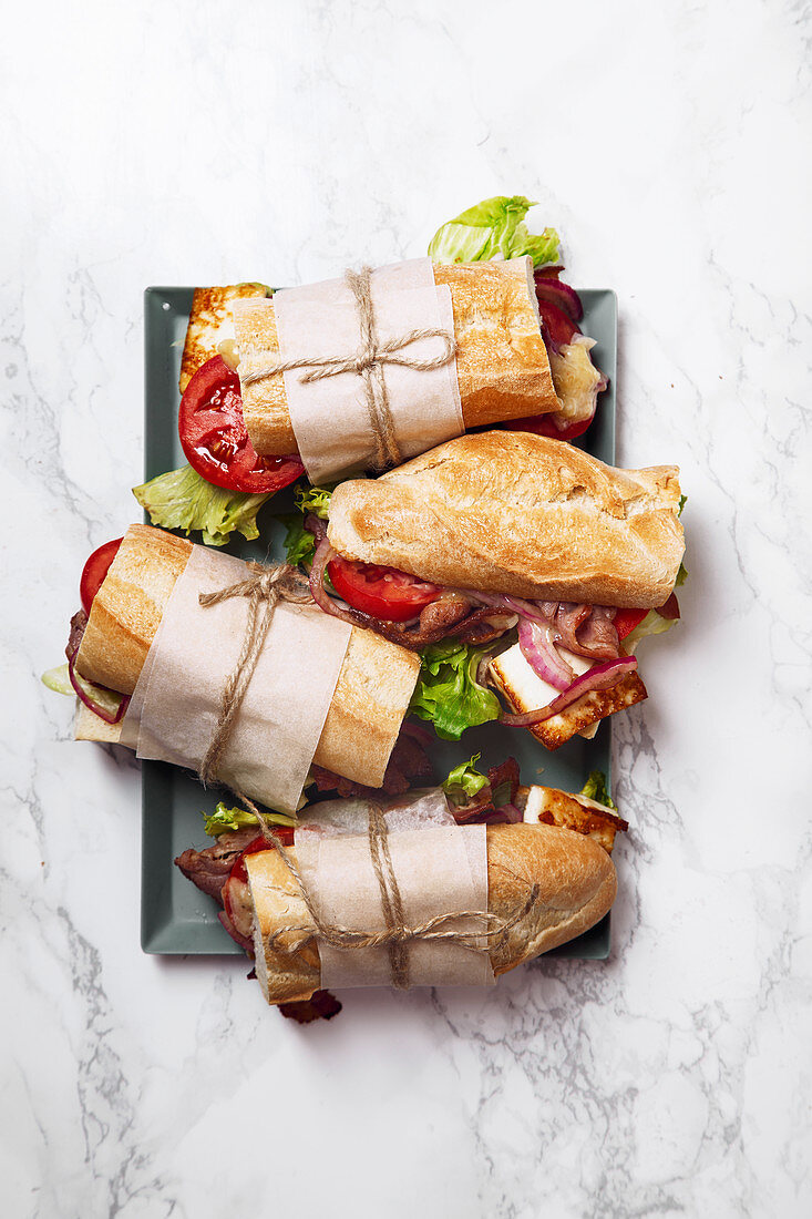 Fresh baguette sandwich bahn-mi styled, bacon, roasted cheese, tomatoes and lettuce on metallic tray on white marble background