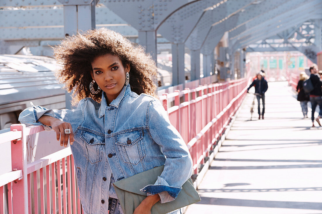 A dark-haired woman wearing jeans and a denim jacket standing on a bridge