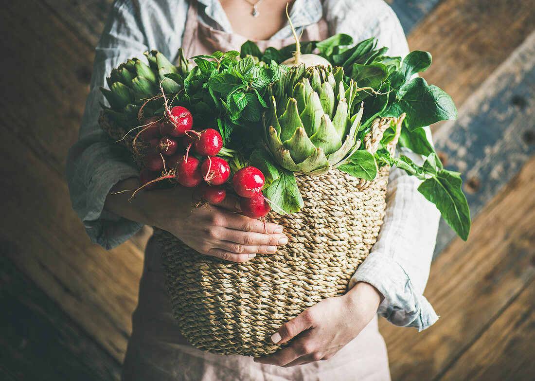 Female farmer in linen apron holding basket of fresh garden vegetables and greens in her hands