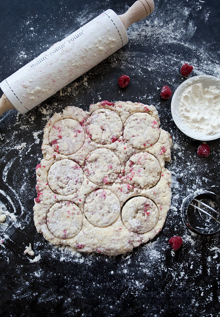 Raspberry scone dough, a rolling pin, dough cutter, flour and raspberries on a black slate surface
