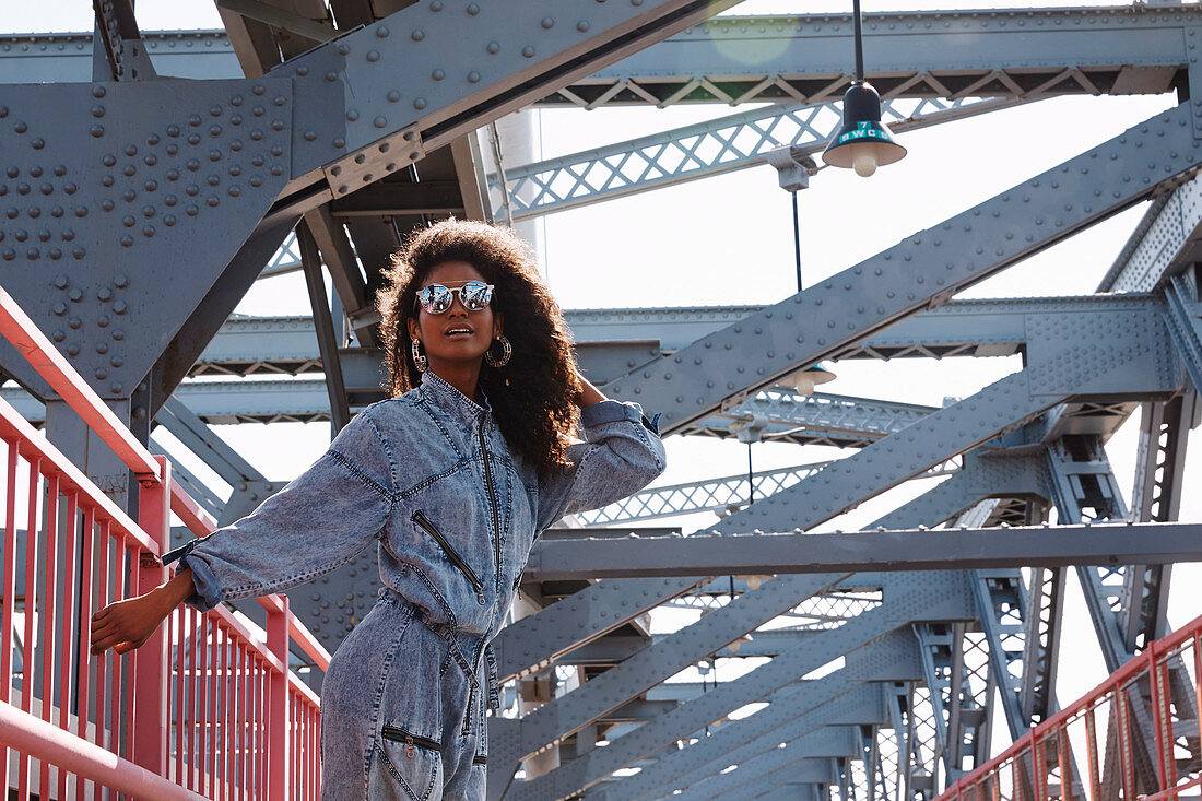 A dark-haired woman wearing sunglasses and a denim jumpsuit on a bridge