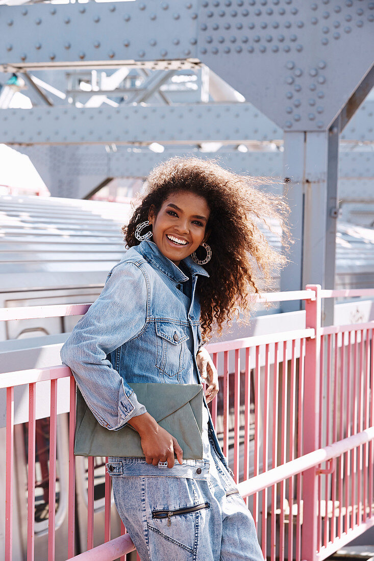 A dark-haired woman wearing jeans and a denim jacket standing on a bridge