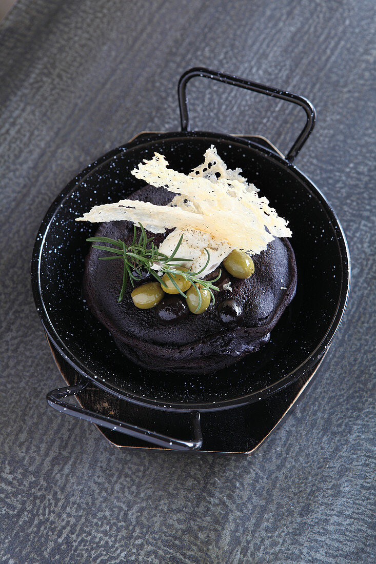 A black sepia pancake decorated with olives and bread chips