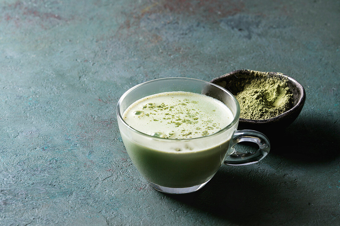 Cup of matcha latte with bowl of matcha tea over grey green texture background