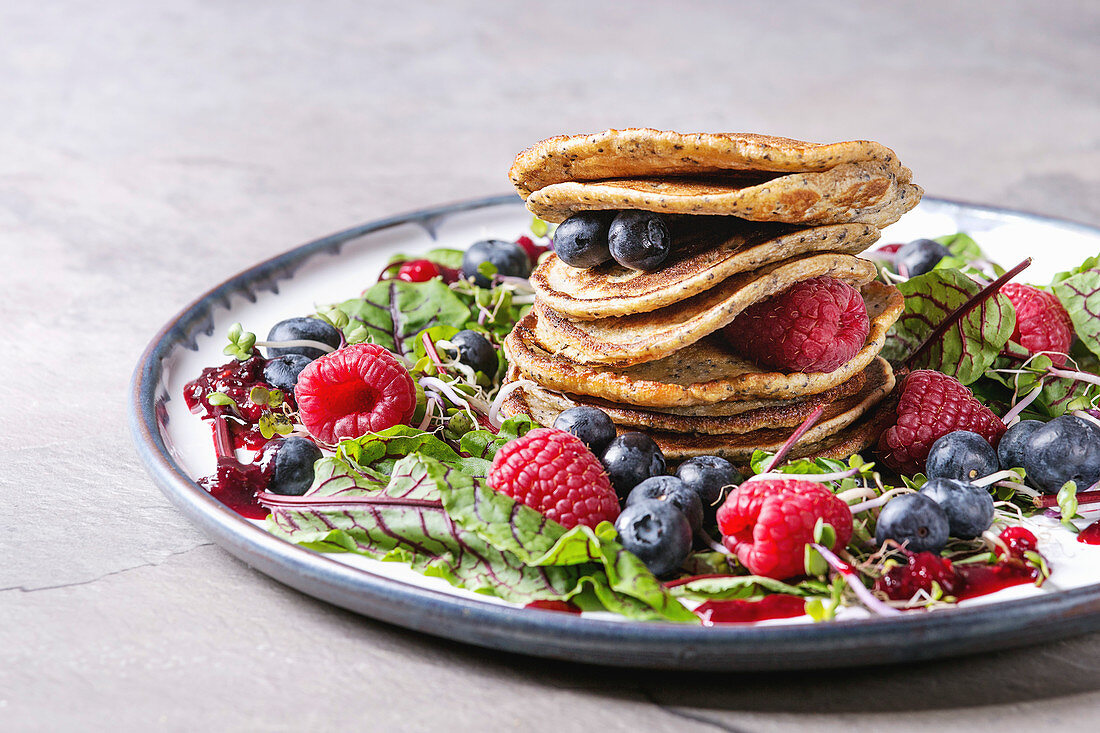 Vegan chickpea pancakes served in plate with green salad young beetroot leaves, sprouts, berries, berry sauce over grey kitchen table