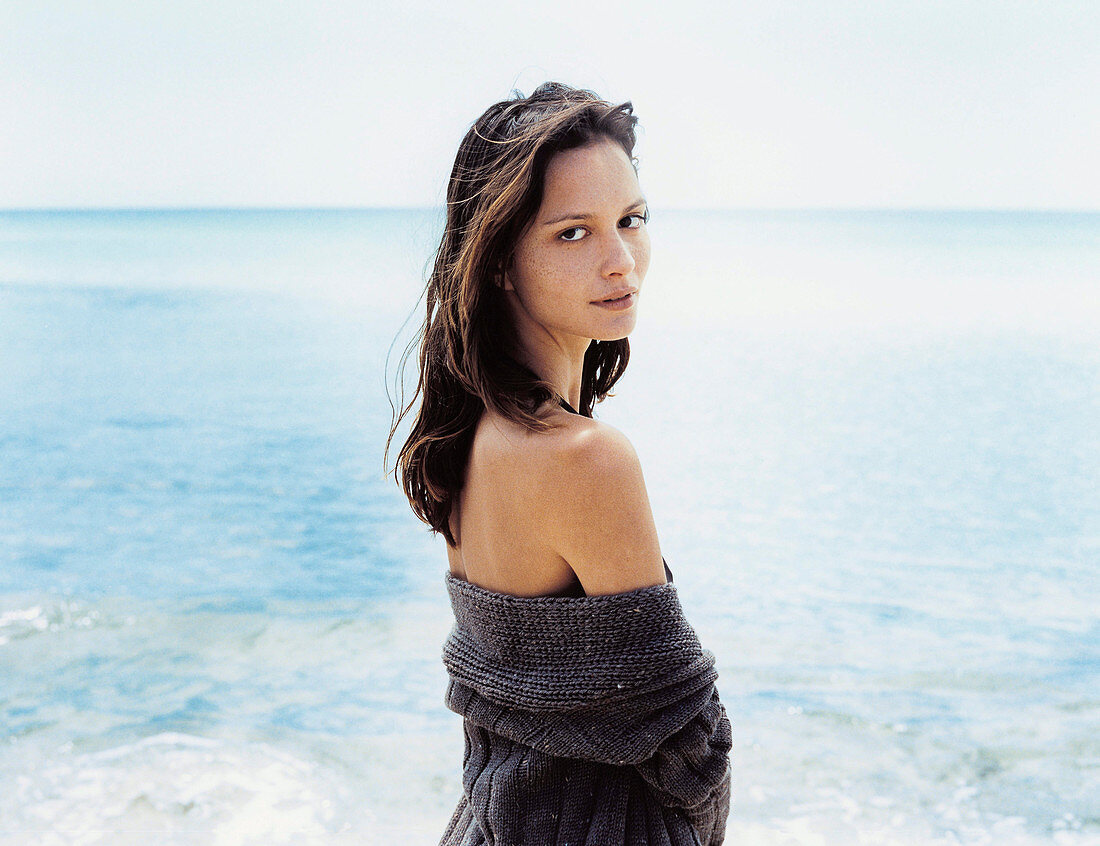 A young woman wearing a grey, off-the-shoulder jumper on a beach