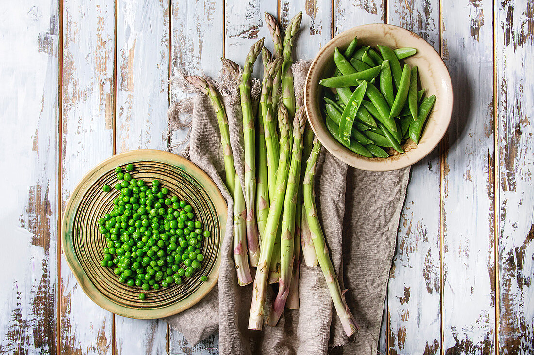 Variety of raw uncooked organic young green vegetables asparagus, peas, pod pea in ceramic plates on linen cloth over white wooden plank background