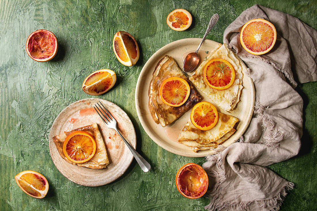 Homemade crepes pancakes served in ceramic plates with bloody oranges and rosemary syrup with sliced sicilian red oranges over green texture background