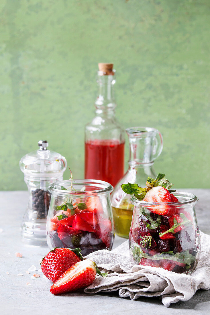 Beetroot and strawberry salad served with arugula and nuts in glass jars with cloth, pepper and bottles of fruit ocet and olive oil over grey table with green wall as background