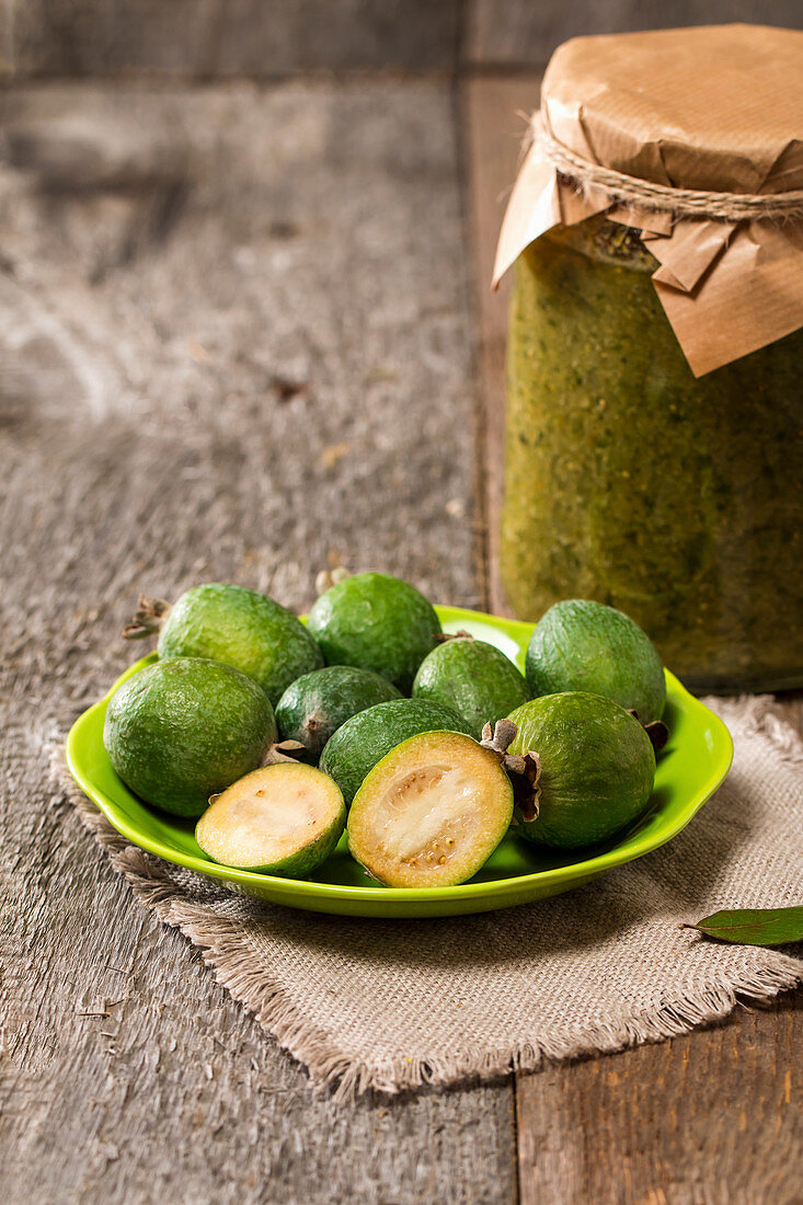 Frische Feijoa auf Teller und ein Glas Feijoa-Marmelade auf Holztisch