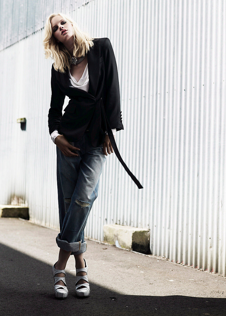 Junge Frau in schwarzem Blazer und hochgekrempelten Jeans vor Wellblechfassade