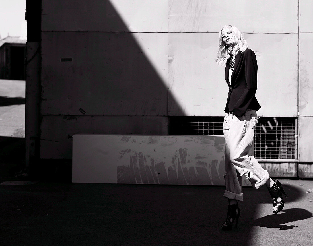 Junge Frau in schwarzem Blazer und hochgekrempelten Jeans vor Industriegebäude (s-w-Aufnahme)
