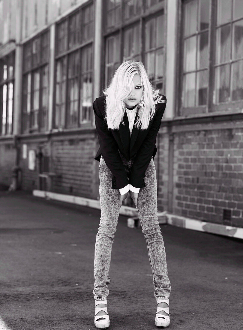 A blonde woman wearing a black jacket and jeans in front of an industrial building (black-and-white shot)