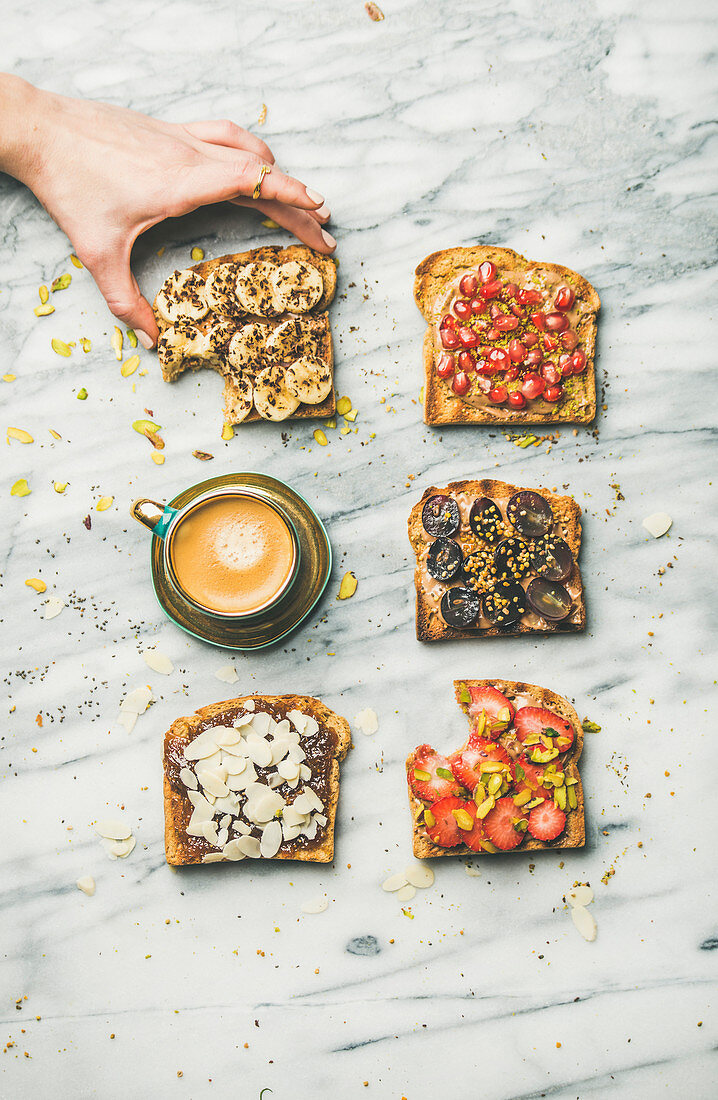 Vegan wholegrain toasts with fruit, seeds, nuts, peanut butter, cup of espresso and woman hand over marble background