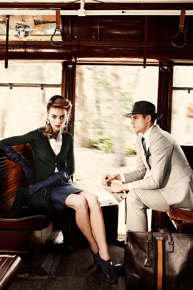 A young woman wearing a black suit and a young man wearing a hat and a light suit sitting on a train