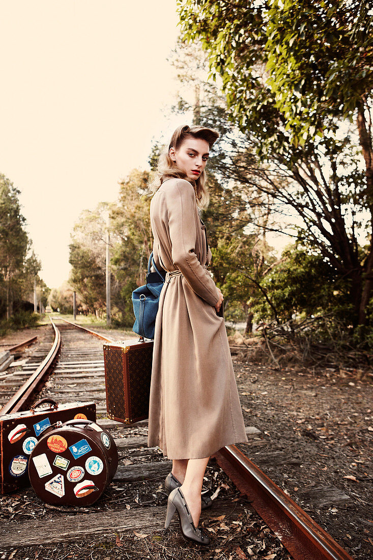 A young woman wearing a long coat standing on railway tracks with suitcases