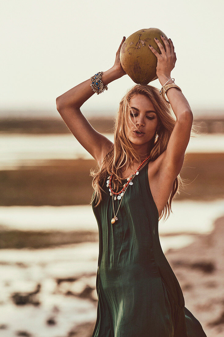 Blonde Frau mit exotischer Frucht über dem Kopf haltend, in grünem Kleid am Strand