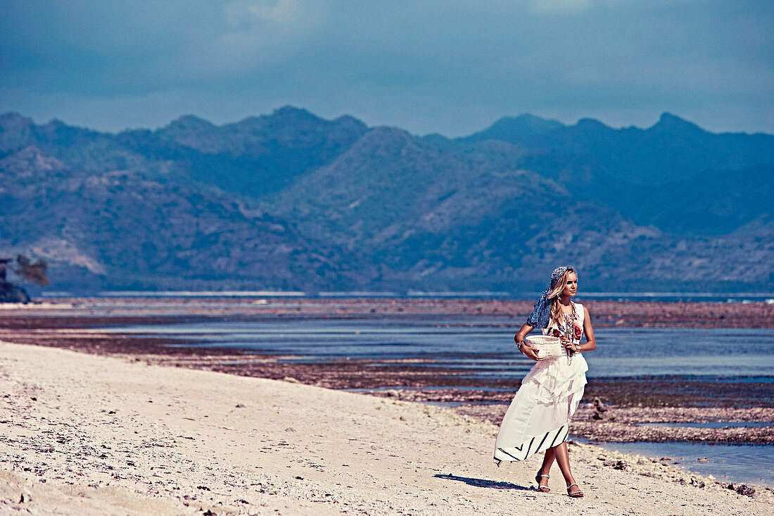 A blonde woman wearing a headscarf and a summer dress on a beach
