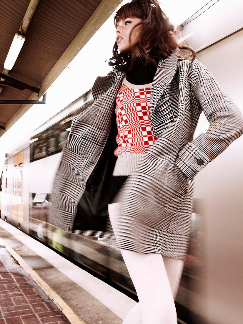 A brunette woman wearing a knitted sleeveless jumper, a black-and-white checked coat and white trousers at a station
