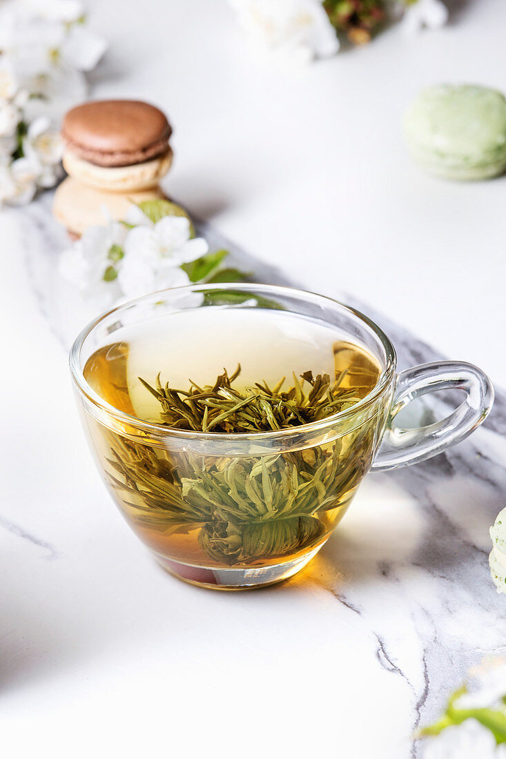 Grüner Tee mit Teeblume in Glastasse, dahinter Macarons und Blüten